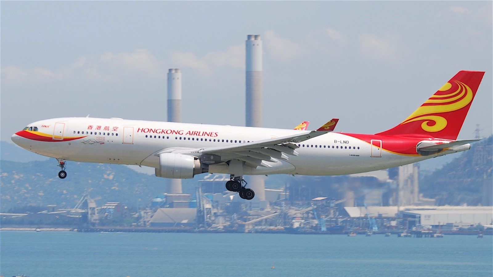 A Hong Kong Airlines airplane is shown in flight near a coastal industrial area, with two tall chimneys visible in the background, suggesting travel disruption due to Typhoon Ragasa.