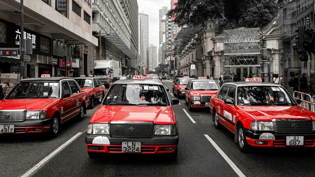 A lineup of red and black taxis with mandatory Journey Recording Systems is parked along a busy urban street in Hong Kong, with tall buildings and storefronts visible in the background.