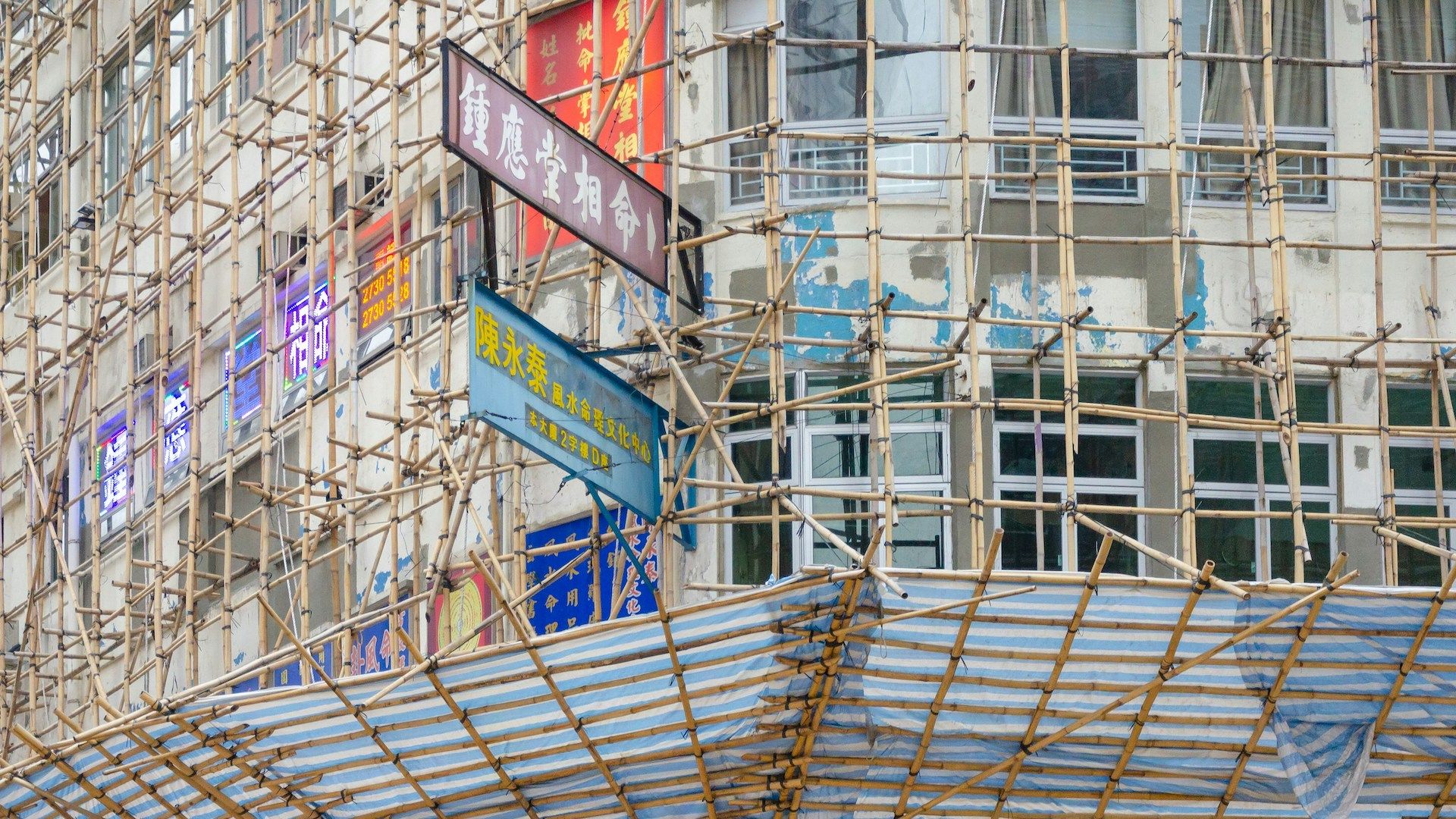 Bamboo and metal scaffolding cover the exterior of a building under construction or renovation in Hong Kong, with signs and windows visible in the background.